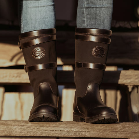 Brown leather boots with a brand logo worn by a person on wooden steps.